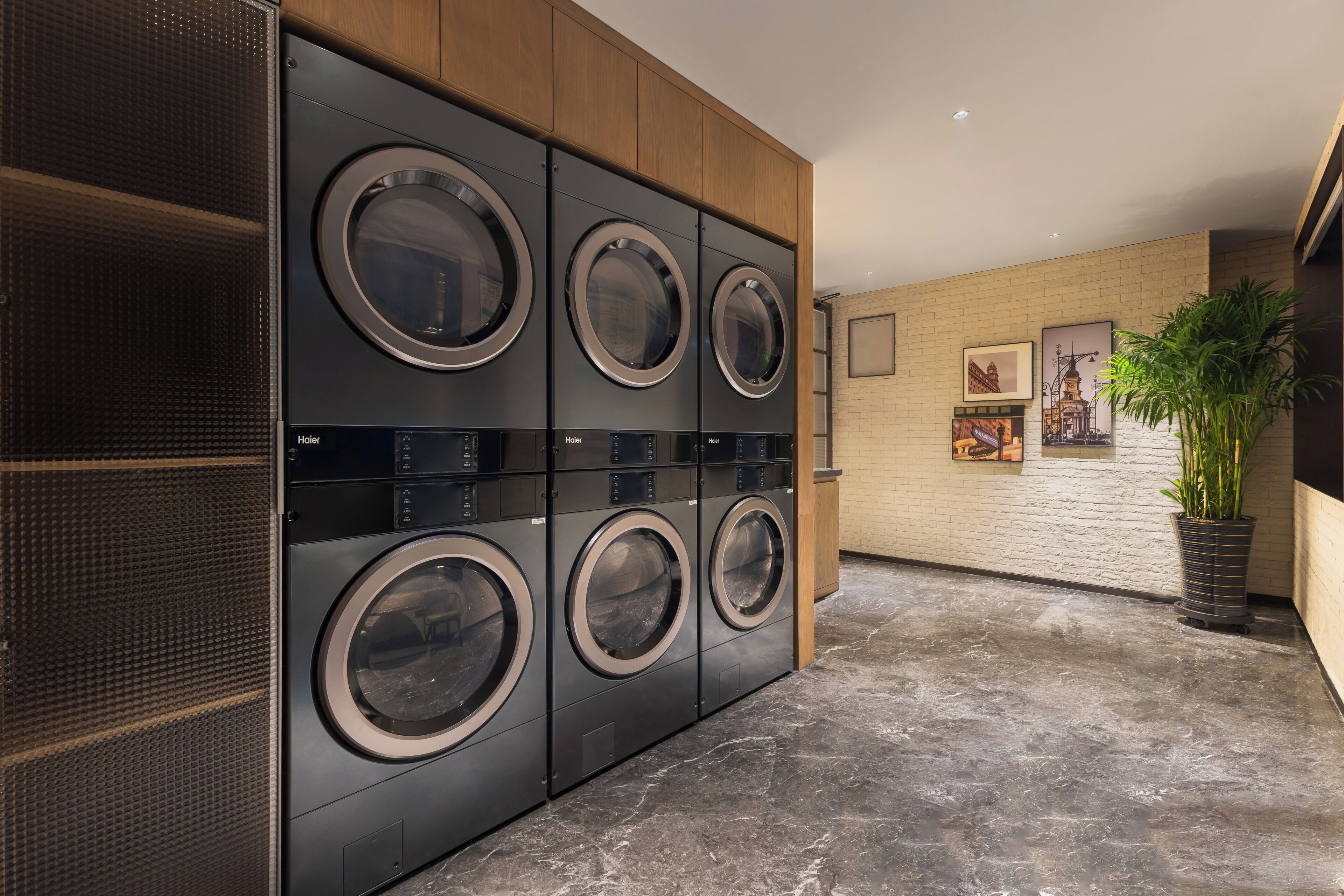 A laundry room with multiple front-loading washers and dryers, a potted plant, and a brick wall with framed pictures.