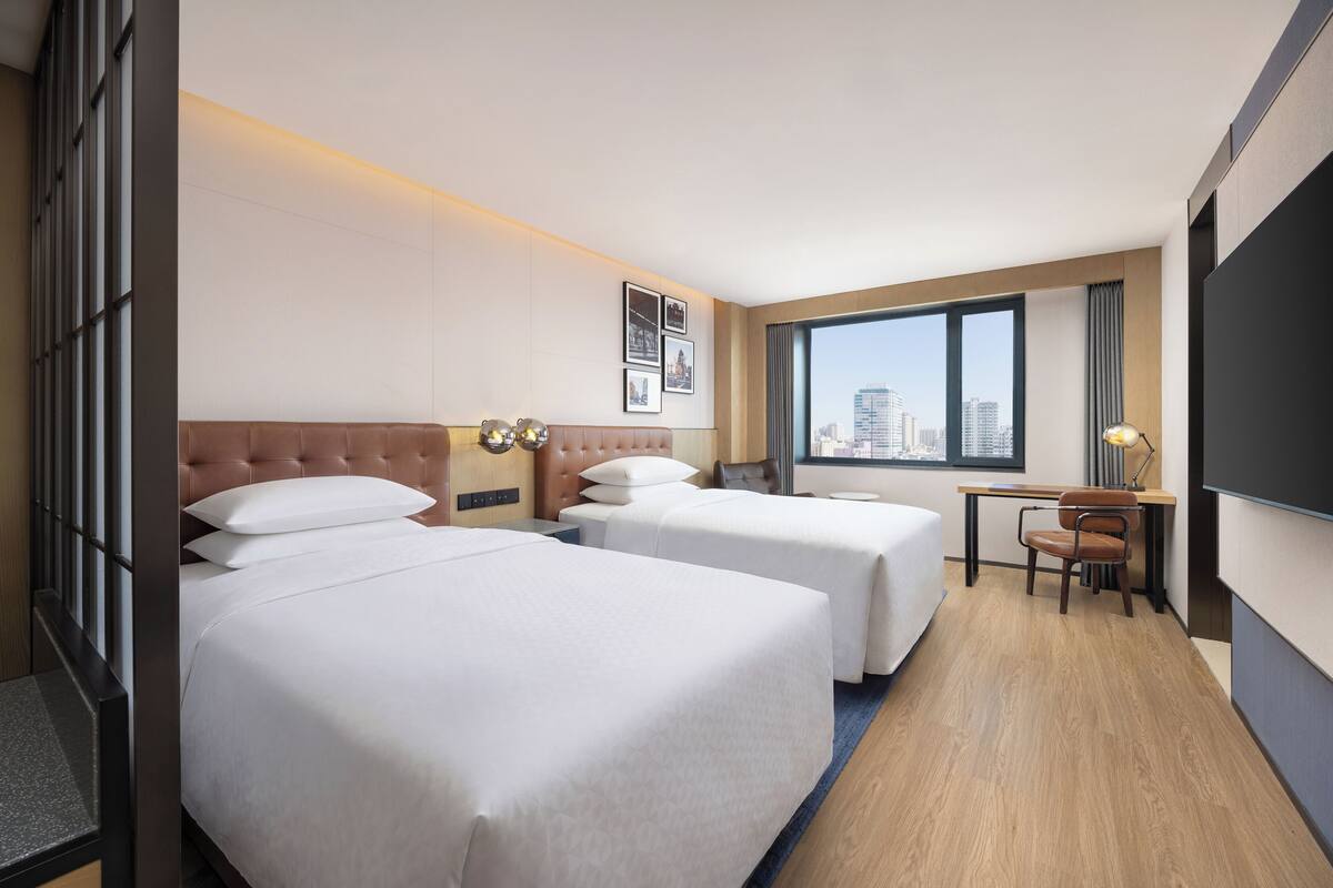 A hotel room with two beds, a desk, a chair, a large window, and a city view.