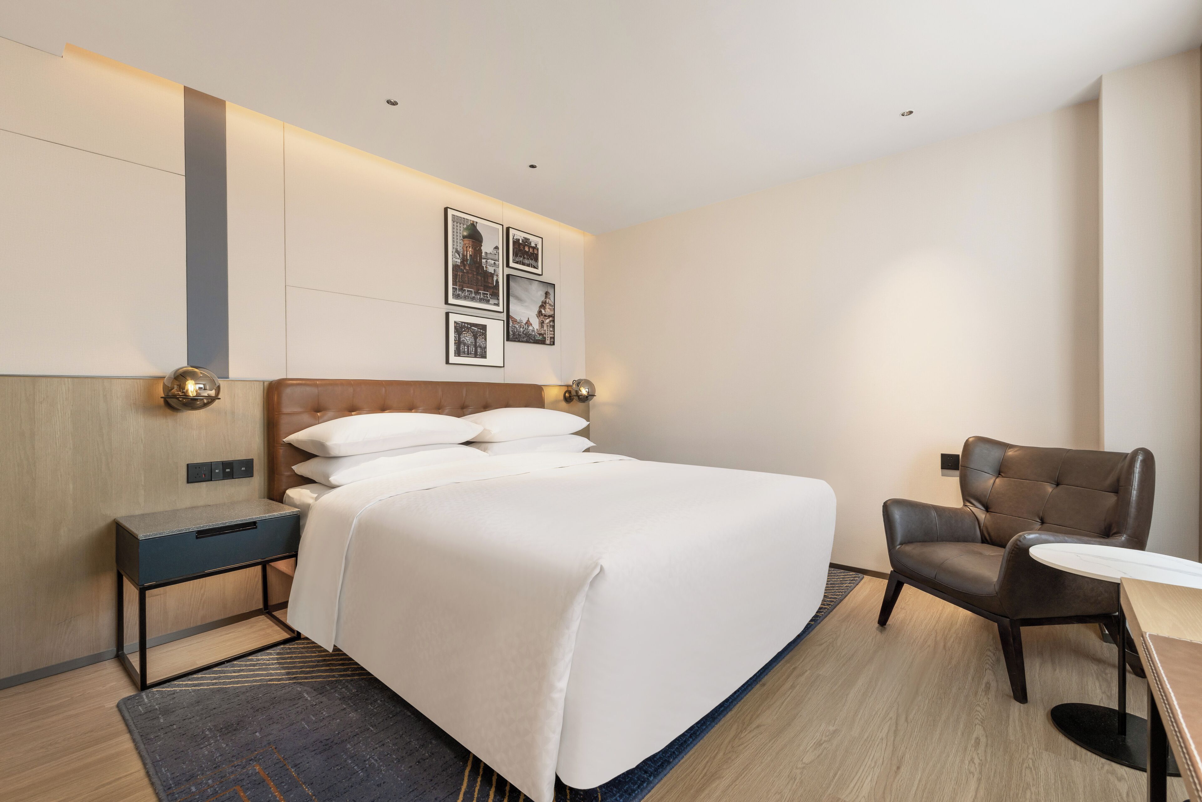 Quarto de hotel com cama grande, mesa de cabeceira, cadeira, mesinha e quadros pendurados na parede.