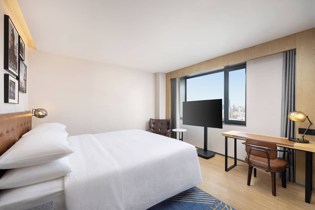 A hotel room with a bed, a desk, a chair, a TV, and a window with a city view.