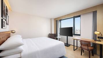 A hotel room with a bed, a desk, a chair, a TV, and a window with a city view.