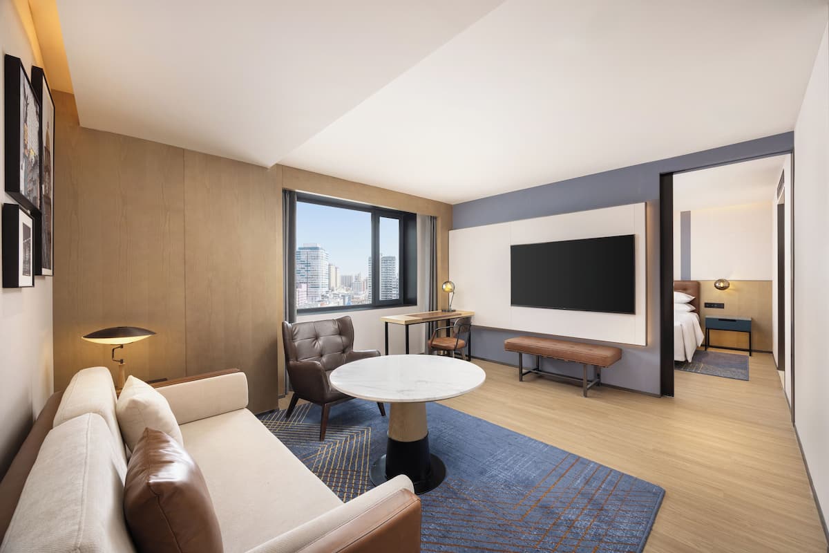 A modern hotel room with a sofa, a round table, a chair, a TV, and a city view.