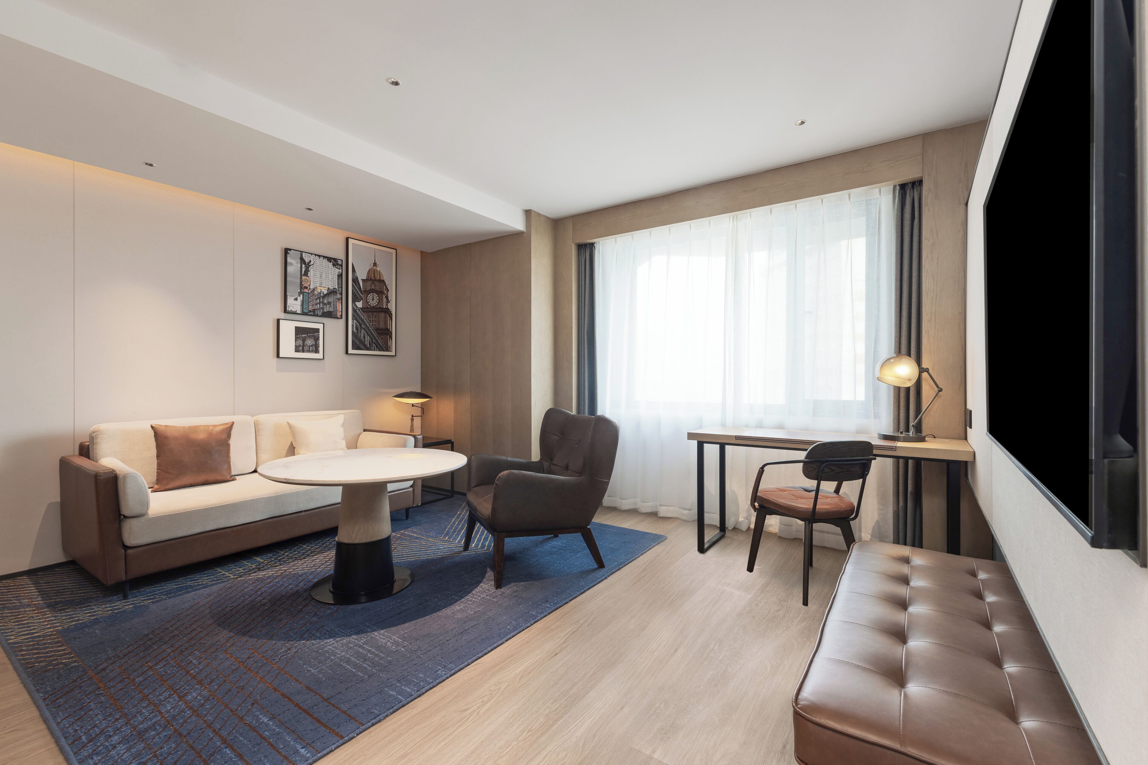 A modern hotel room with a sofa, a round table, a chair, a desk, and a large window.