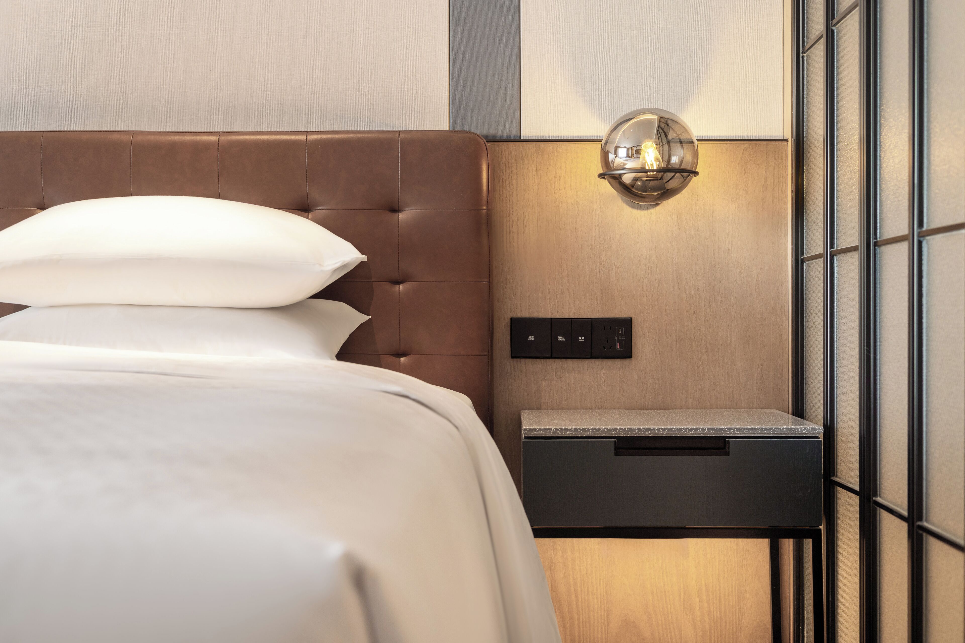 A neatly made bed with a headboard, a bedside table with a lamp, and a wall-mounted switchboard.