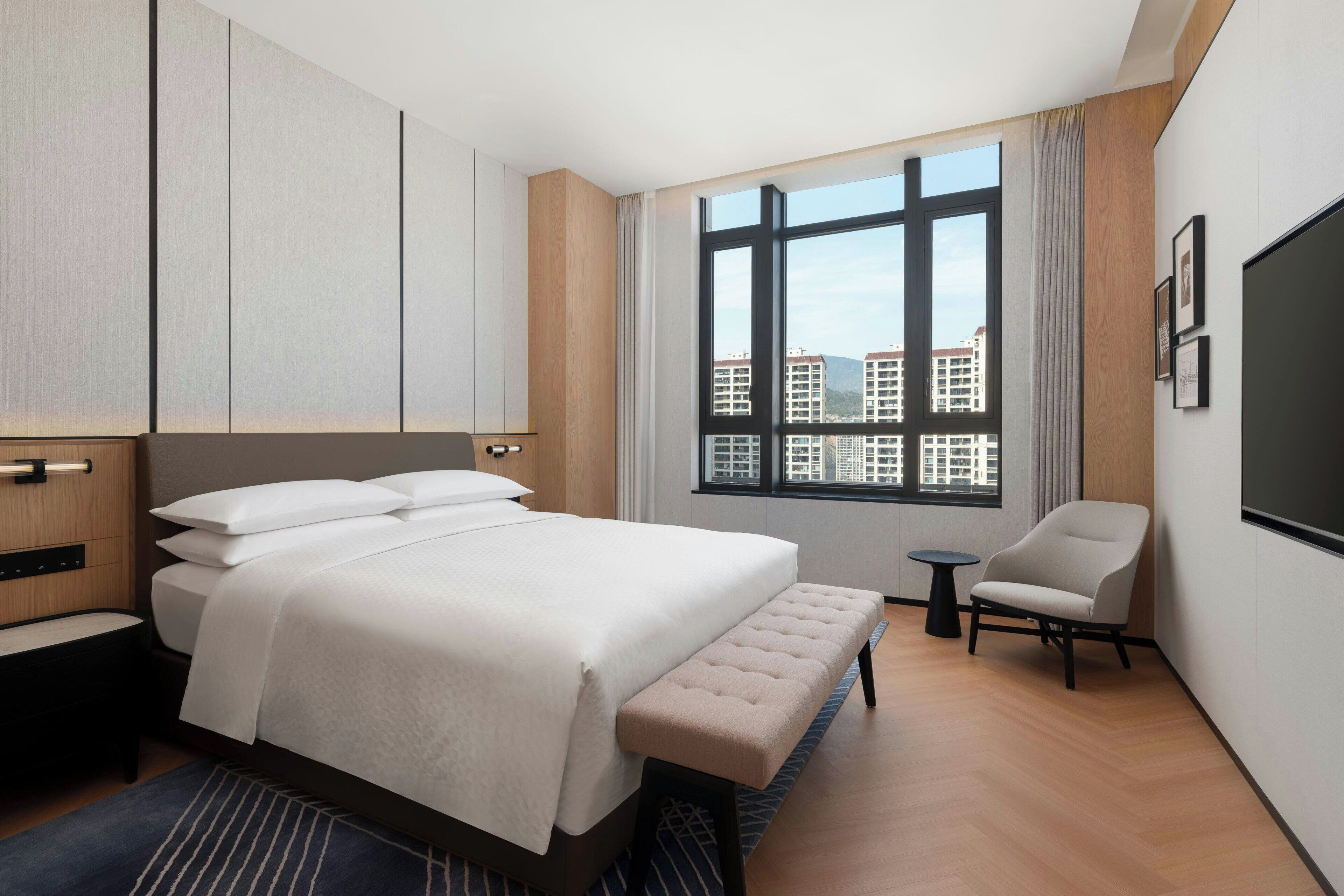 Ein modernes Hotelzimmer mit einem großen Bett, einem kleinen runden Tisch, einem Sessel und einem großen Fenster mit Blick auf die Stadt.