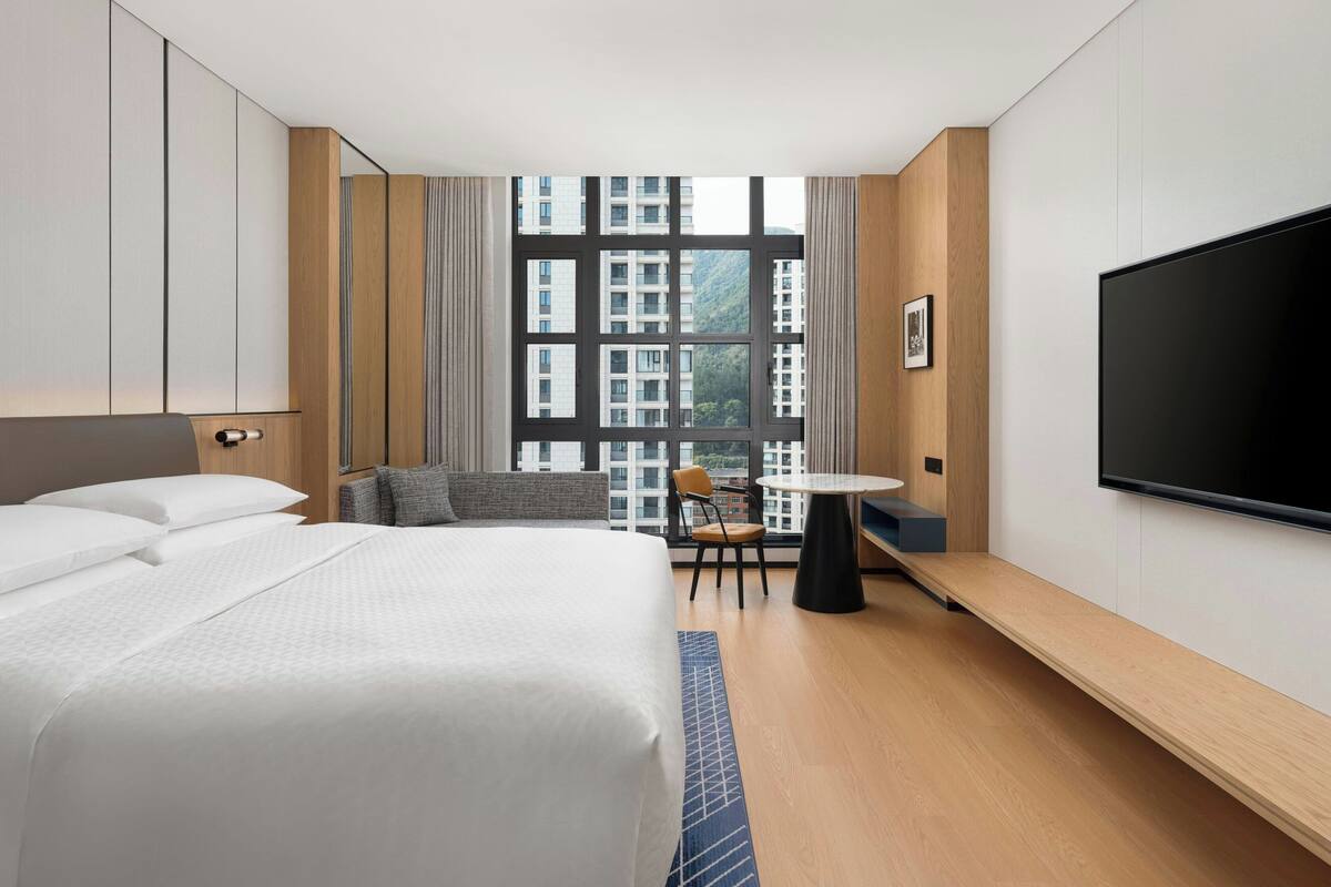 A hotel room with a bed, a TV, a small table, and a view of buildings through large windows.