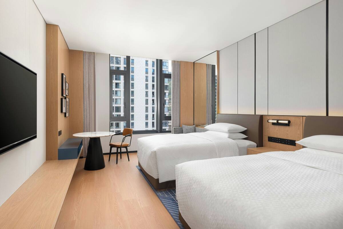 A hotel room with two beds, a desk, a chair, a TV, and a large window with a city view.
