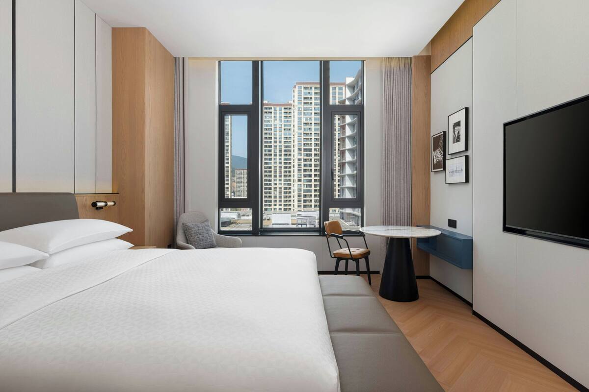A hotel room with a large bed, a desk, a chair, a TV, and a window with a city view.