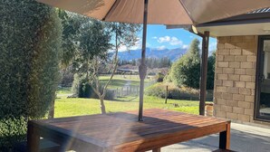 An outdoor patio with a wooden table and bench, a large umbrella, and a view of mountains and greenery. - Stunning views in quiet location, luggate. 10 mins out of Wanaka. (Luggate)