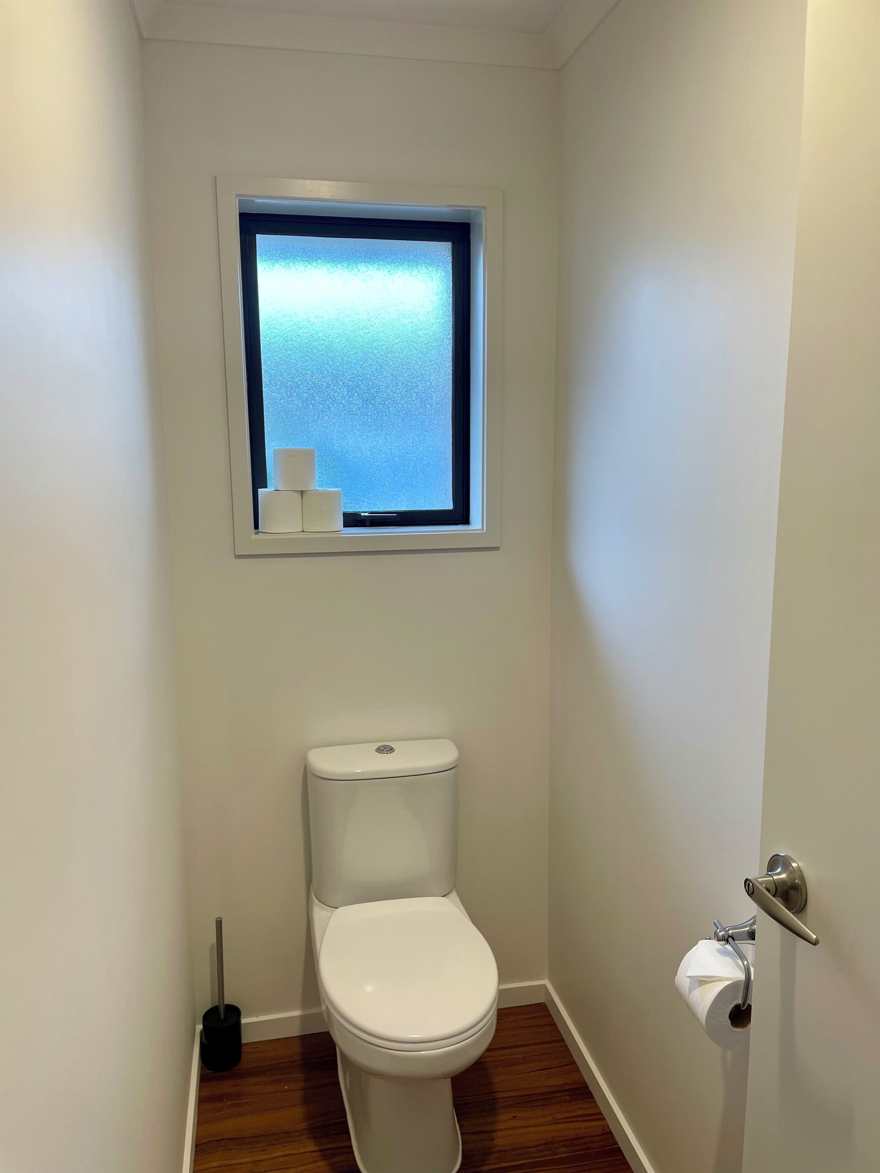 Une petite salle de bain blanche, équipée de toilettes, d’une fenêtre et d’un porte-rouleau de papier toilette.