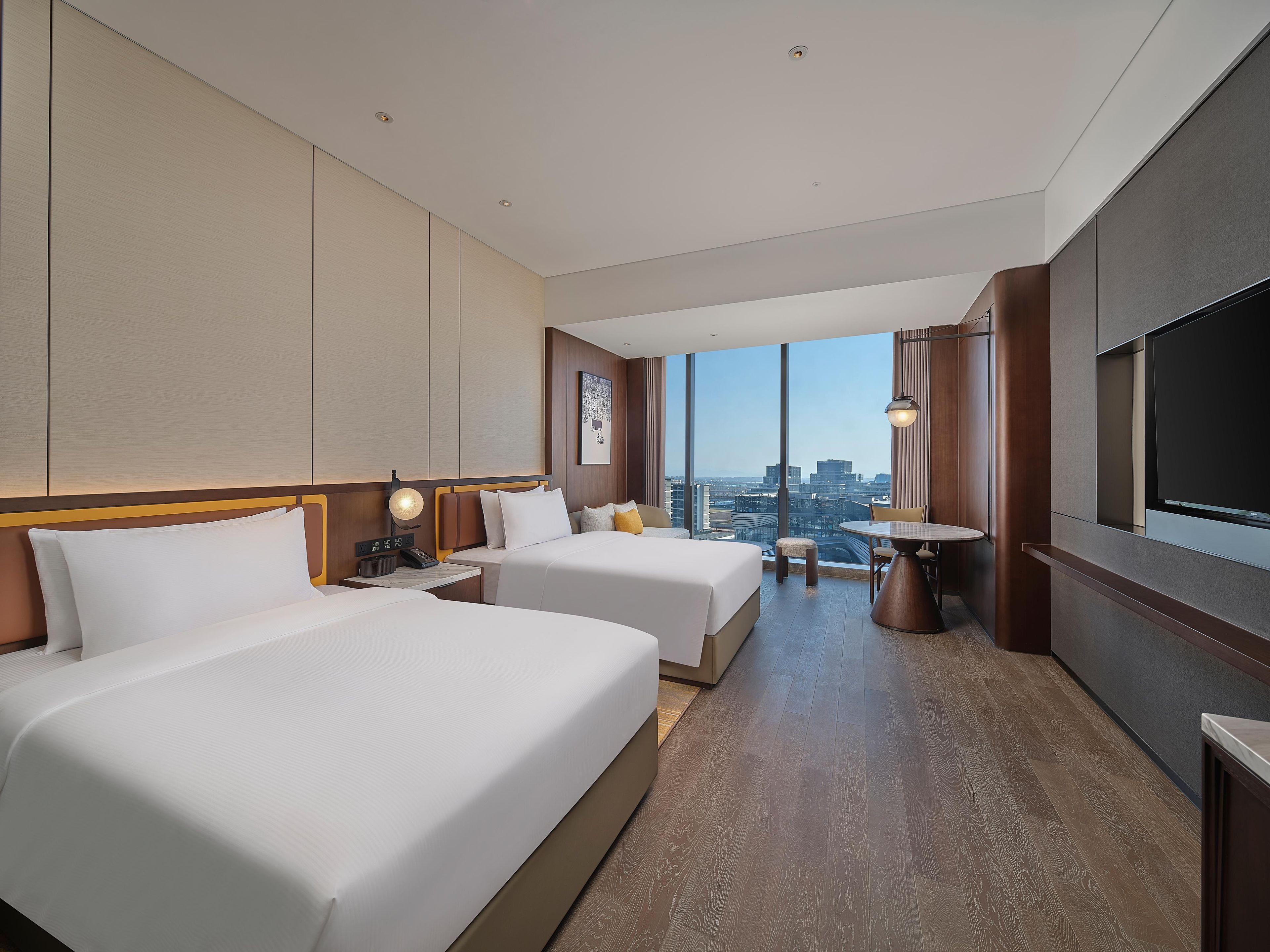 Ein Hotelzimmer mit zwei Betten, einem Esstisch, einem Fernseher und Blick auf die Stadt.
