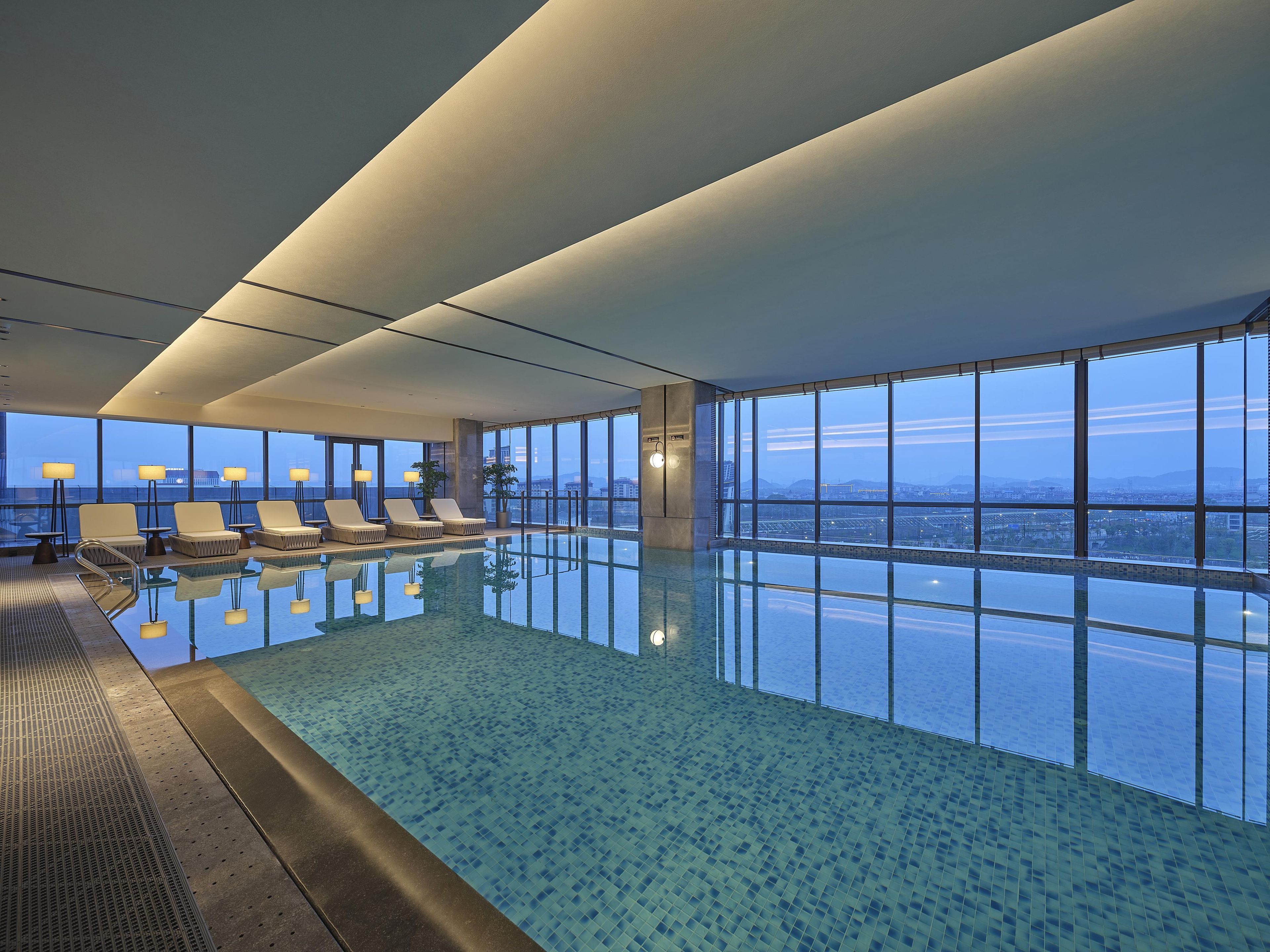 Una amplia piscina cubierta con agua cristalina, rodeada de sillones de descanso y grandes ventanales con vistas a la ciudad.