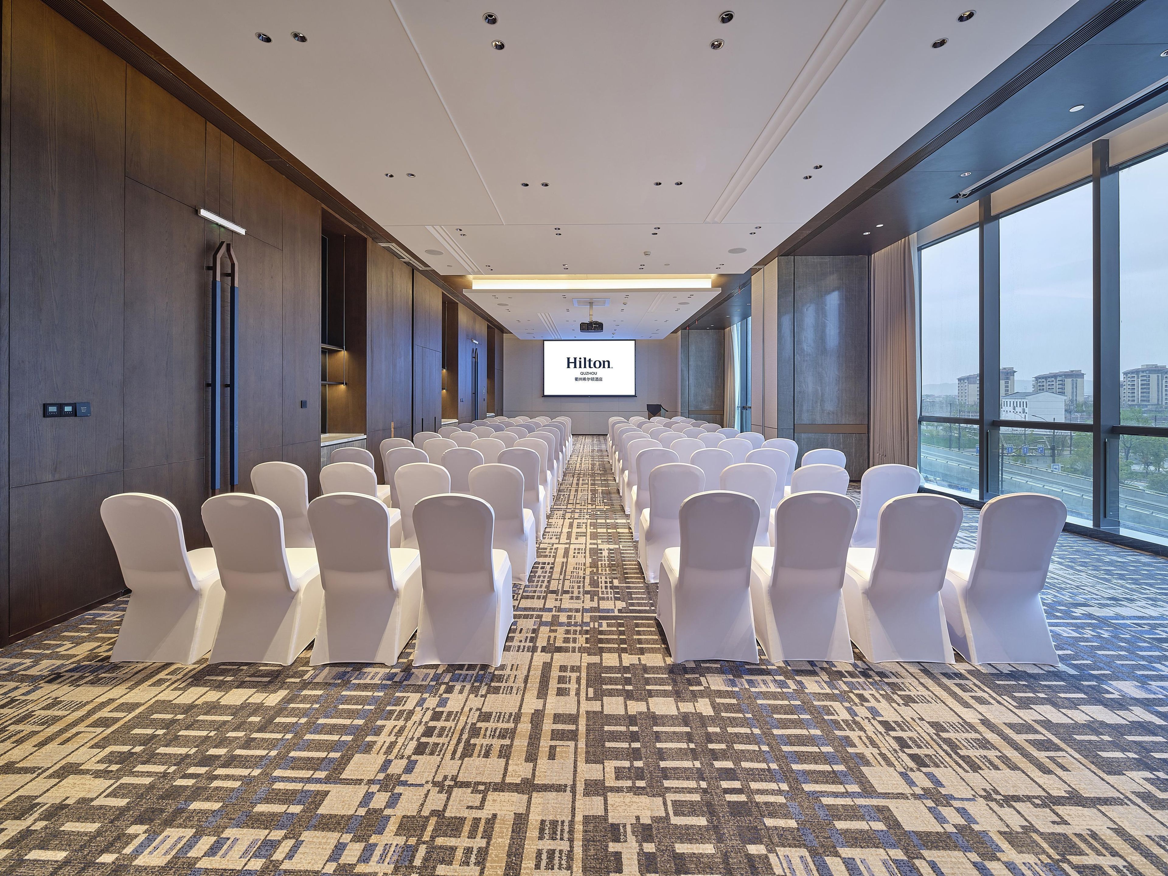 Un salón de conferencias con filas de sillas blancas, una pantalla grande al frente y vistas de la ciudad a través de amplias ventanas.