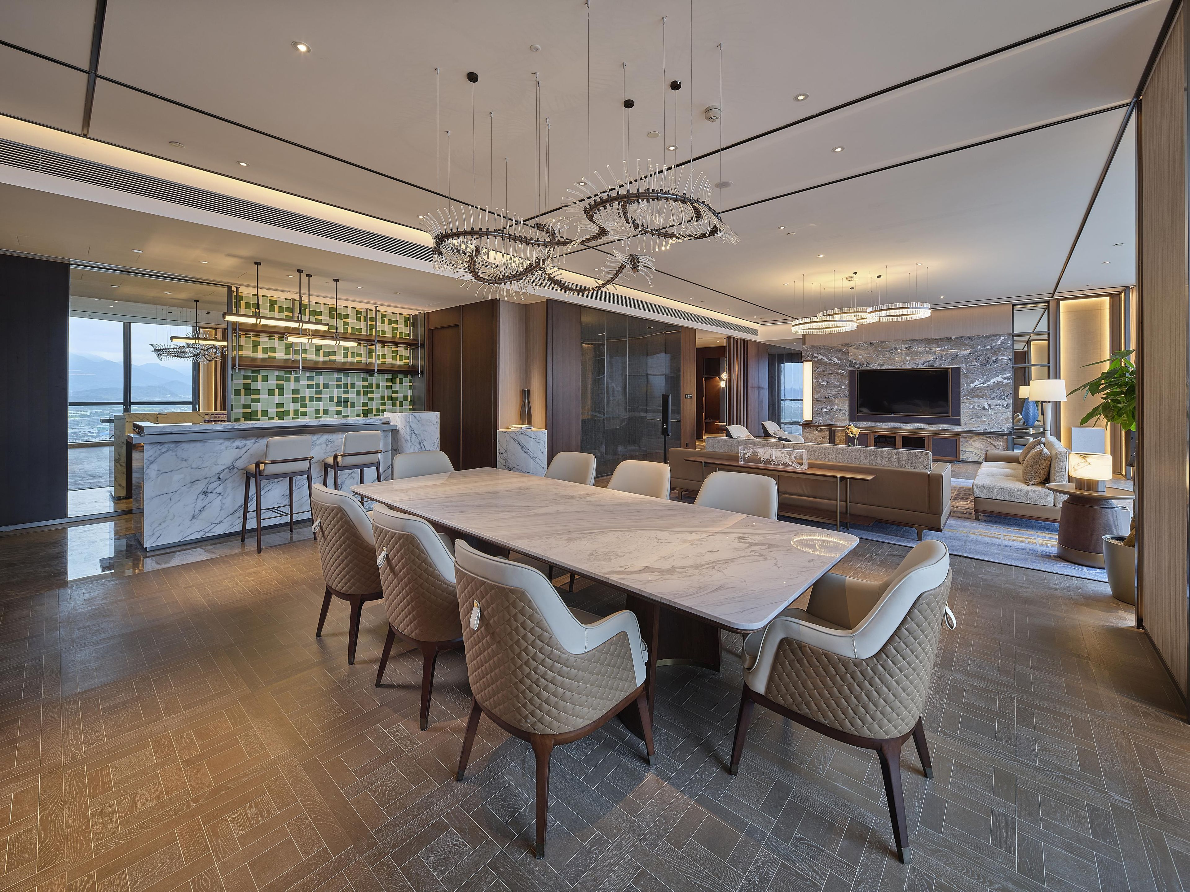 Un interior moderno con zona de comedor que incluye una mesa de mármol y sillas acolchadas, cocina con salpicadero de azulejos verdes y sala de estar con un televisor grande y asientos.