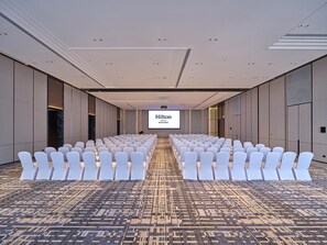A conference room with rows of white chairs, a large screen displaying 'Hilton', and a patterned carpet.