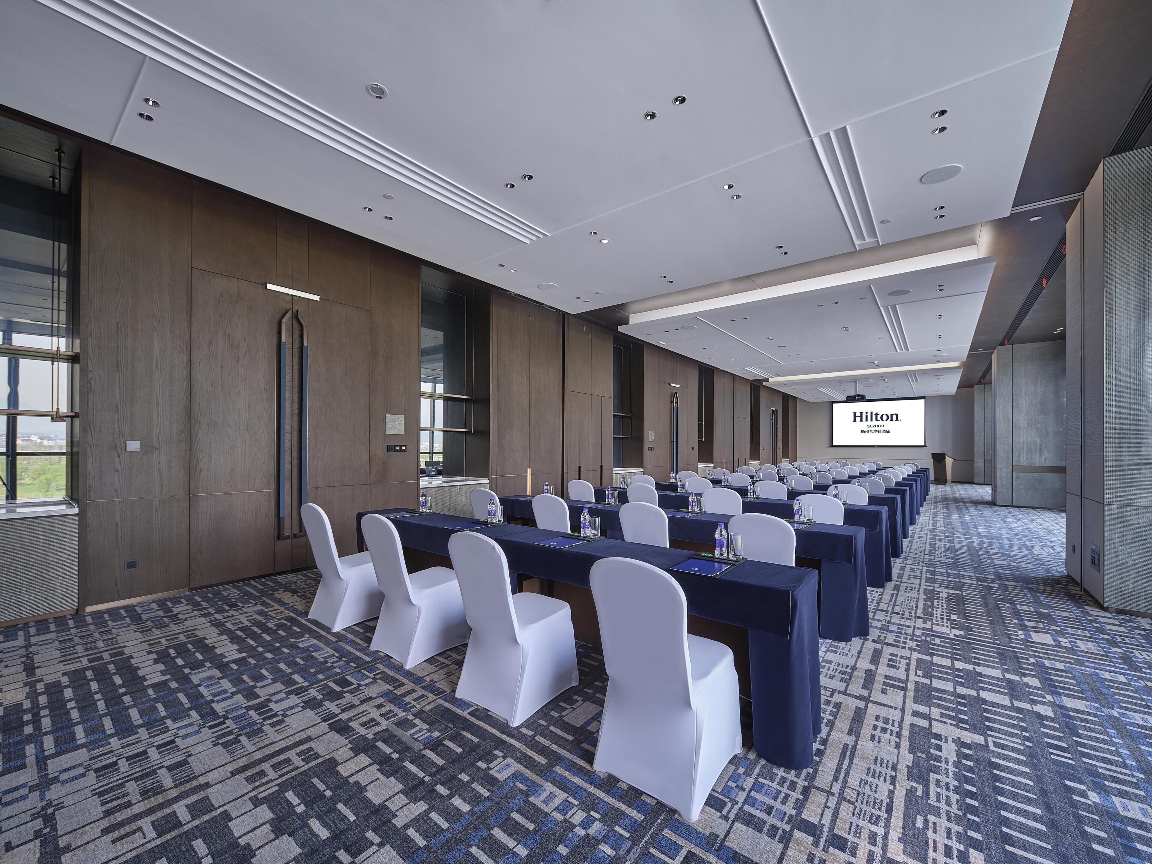 Un salón de conferencias con filas de mesas y sillas, una pantalla grande mostrando 'Hilton' y una alfombra estampada.