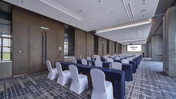 Un salón de conferencias con filas de mesas y sillas, una pantalla grande mostrando 'Hilton' y una alfombra estampada.