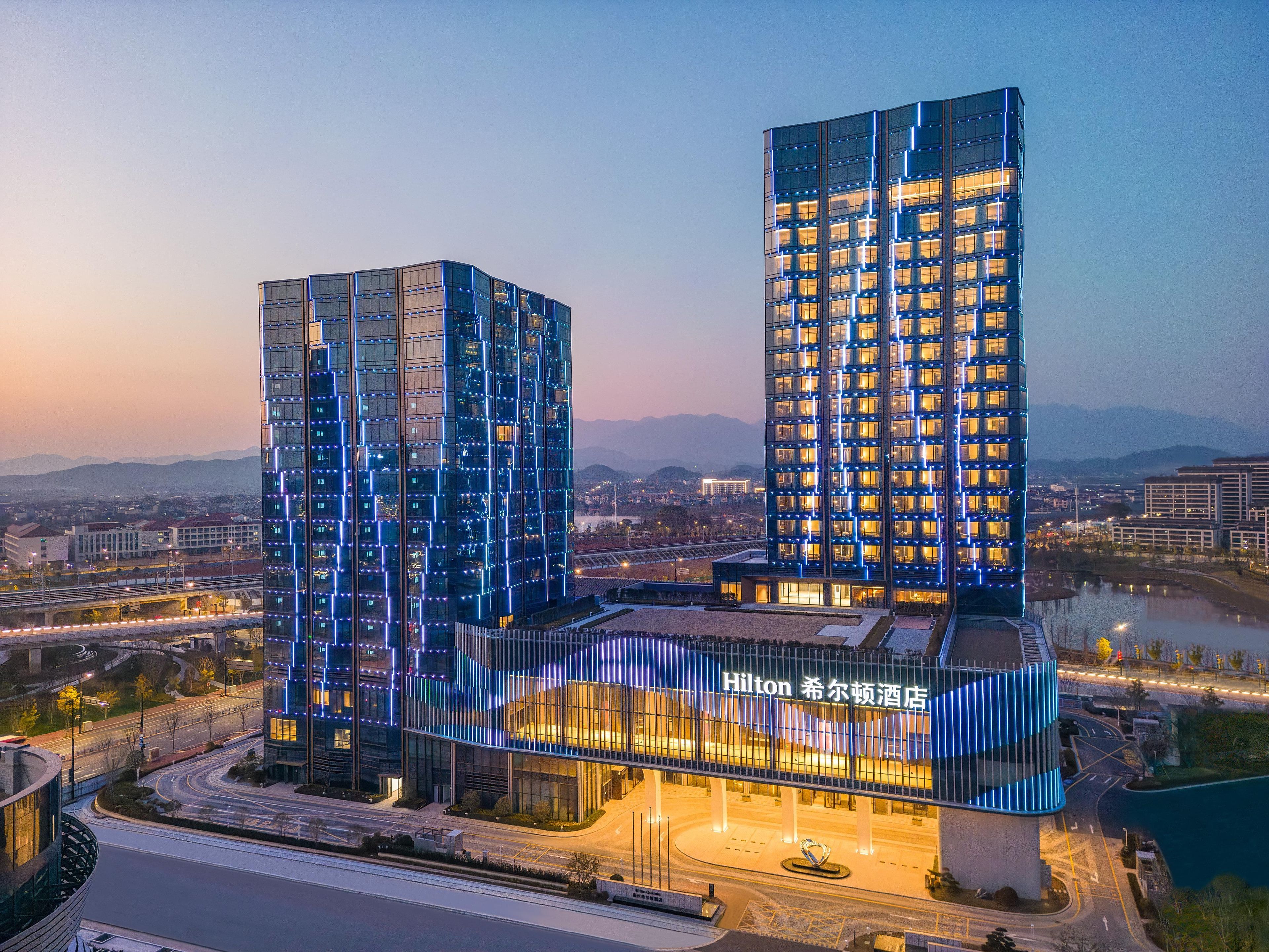 Un hotel moderno con una fachada curva y varios pisos, rodeado por un paisaje urbano y una masa de agua.