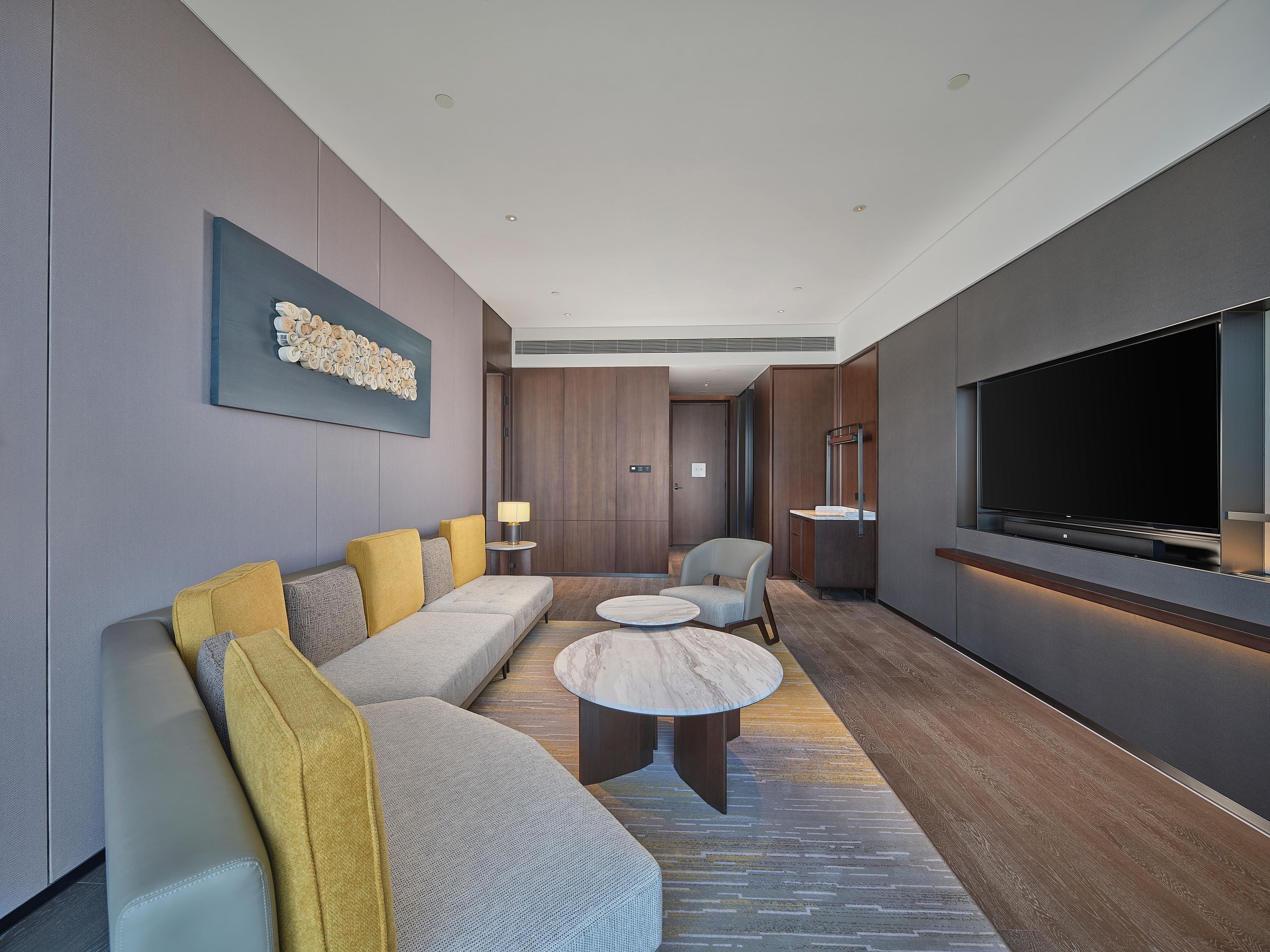 Een moderne hotelkamer met een groot flatscreen televisietoestel, een zithoek, een rond bijzettafeltje en een decoratief wandobject.
