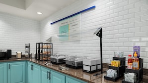 A kitchen with teal cabinets, granite countertops, and a white brick backsplash. There are food dispensers, a coffee machine, and condiments on the counter. - Garner Hotel Madison Huntsville Airport by IHG (Madison)