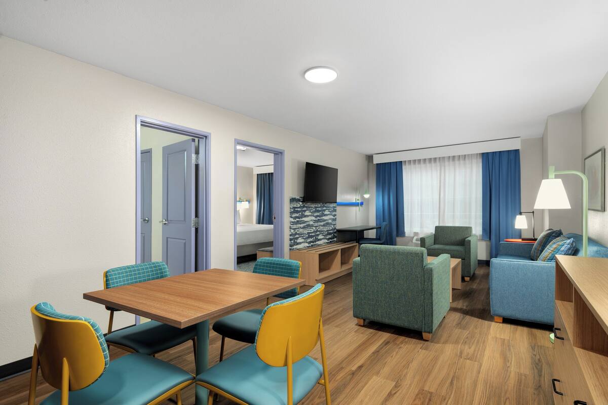A hotel room with a dining table, chairs, a sofa, a TV, and a lamp.