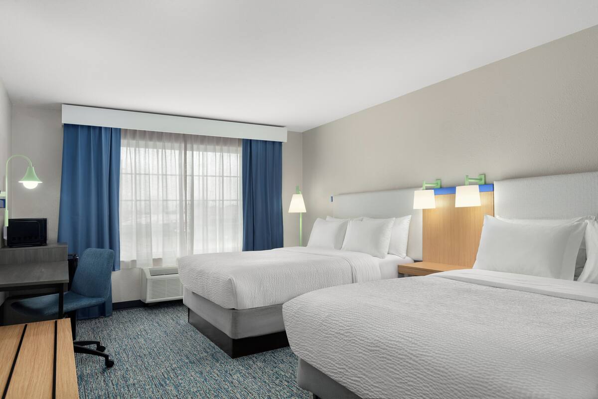 A hotel room with two beds, a desk, a chair, a desk lamp, and a window with curtains.