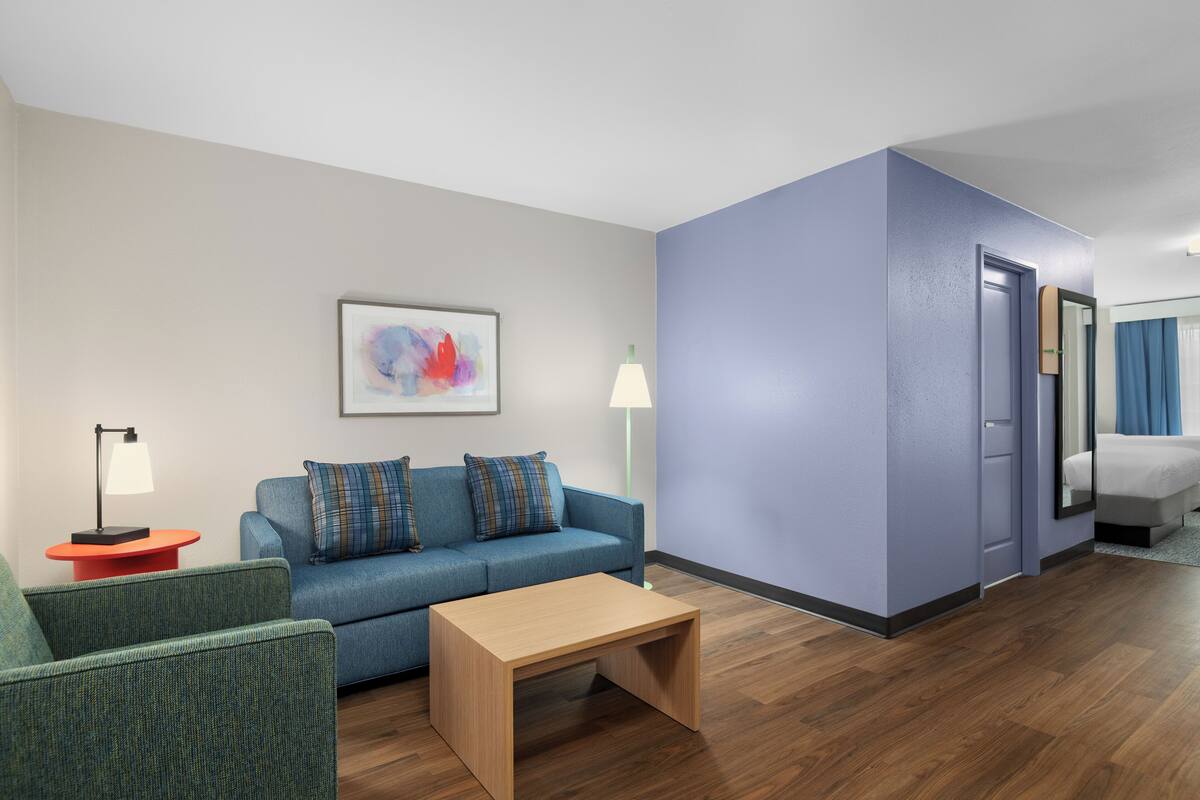 A modern hotel room with a blue sofa, a wooden coffee table, a painting on the wall, and a lamp.