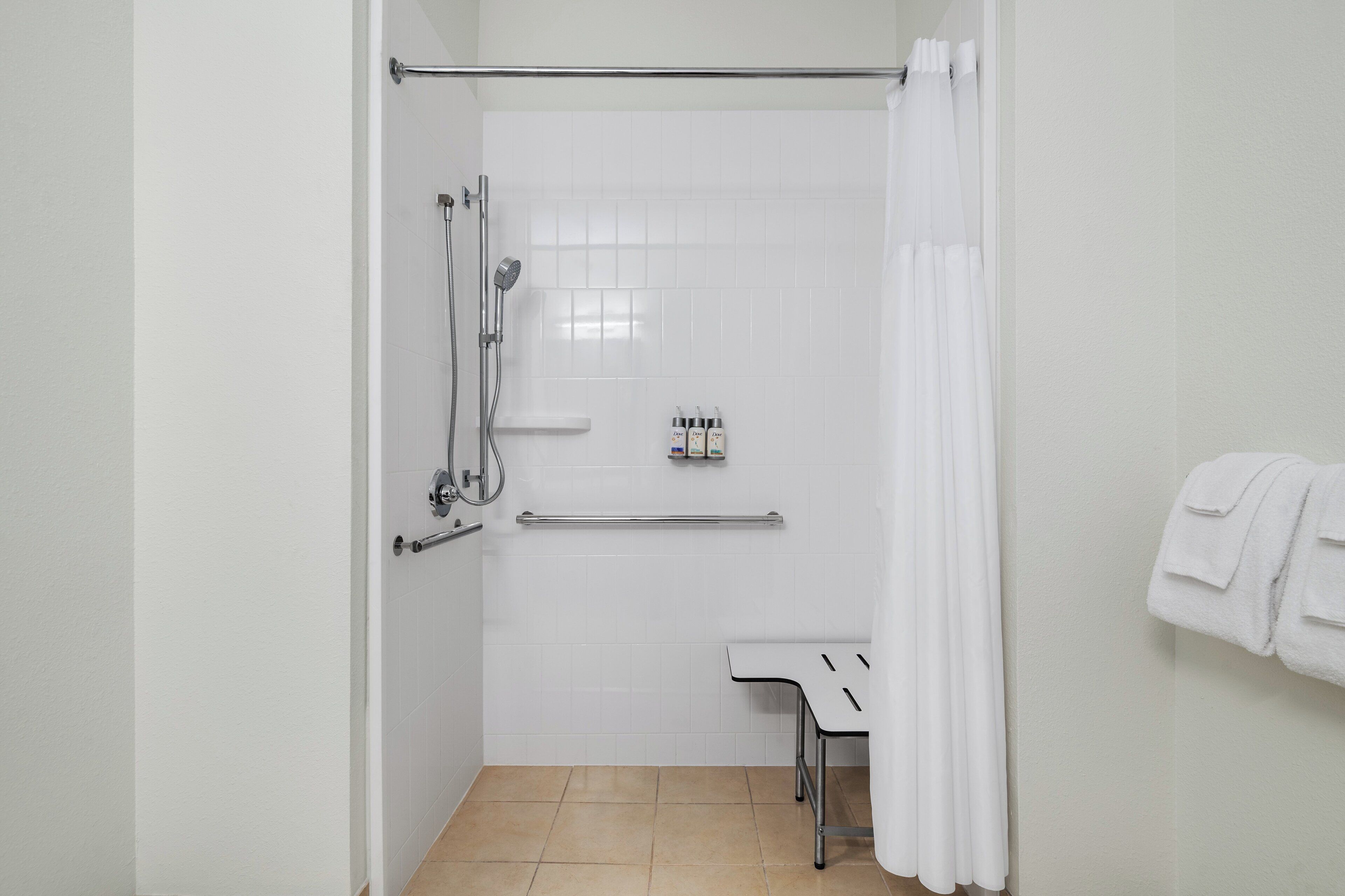 A small, white bathroom with a shower, a towel rack, and a shelf with toiletries.