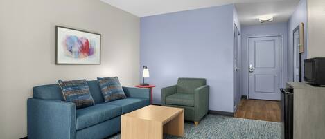 Una habitación con un sofá azul, un sillón verde, una mesita de centro de madera, un cuadro abstracto enmarcado, una mesita con una lámpara y una puerta que da a otra habitación.