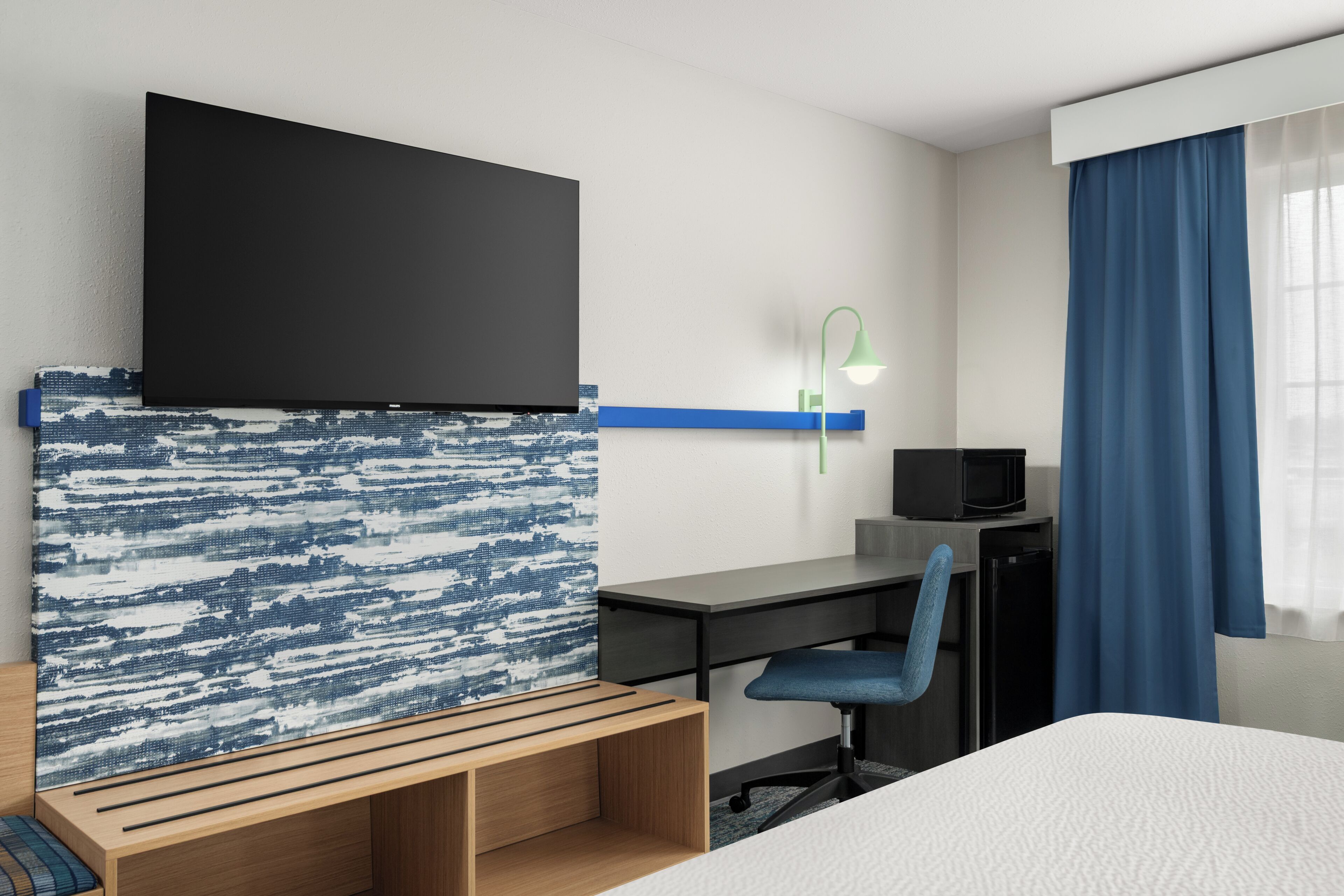 Ein Hotelzimmer mit Bett, Schreibtisch mit Stuhl, Fernseher und Wandleuchte.