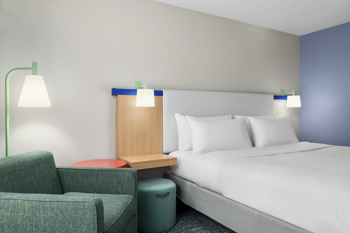 Habitación de hotel con cama, dos lámparas de noche, una mesita de noche, un sofá verde y una pequeña mesa redonda.