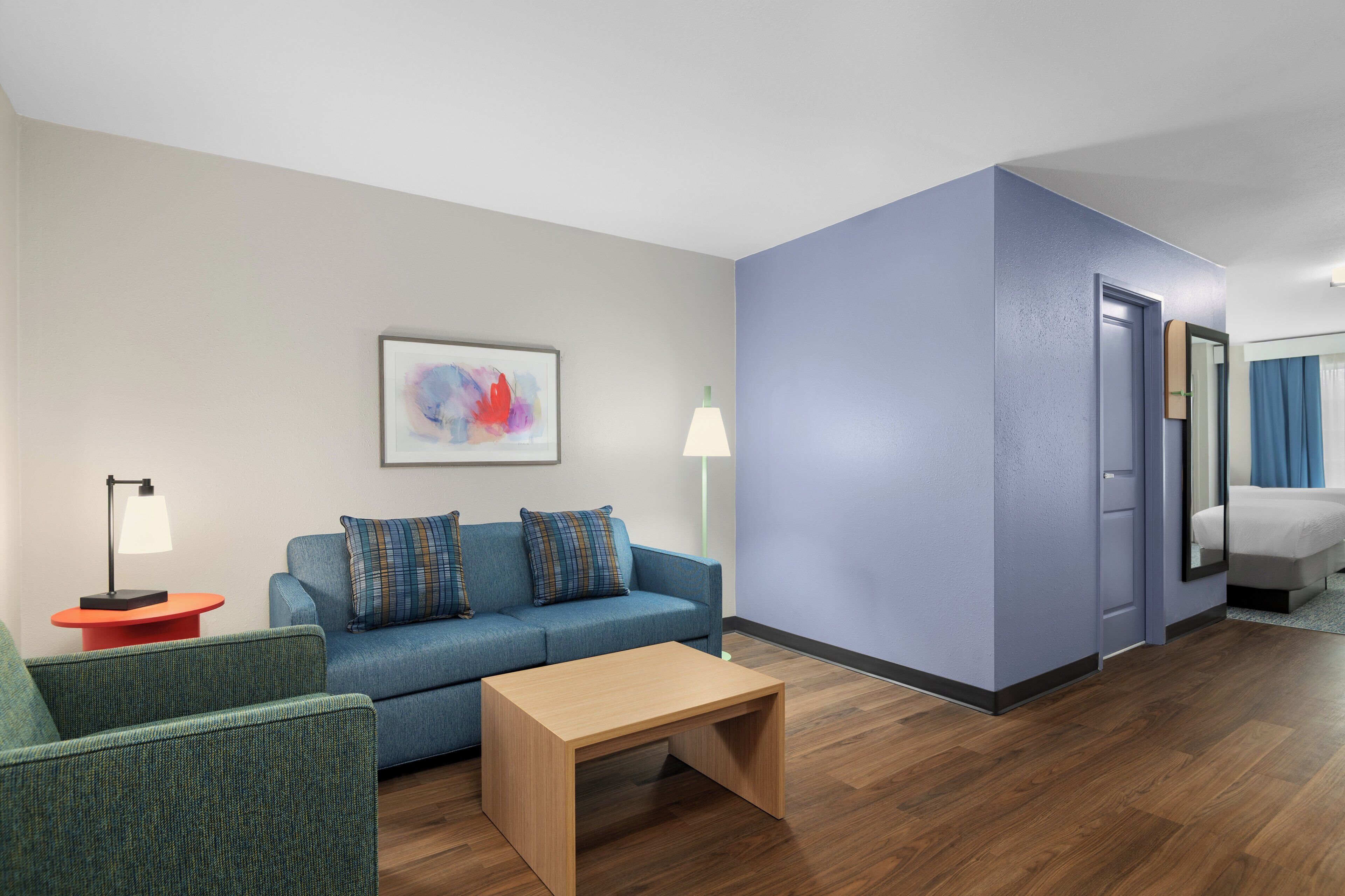 A modern hotel room with a blue sofa, a small wooden table, a floor lamp, and a painting on the wall.
