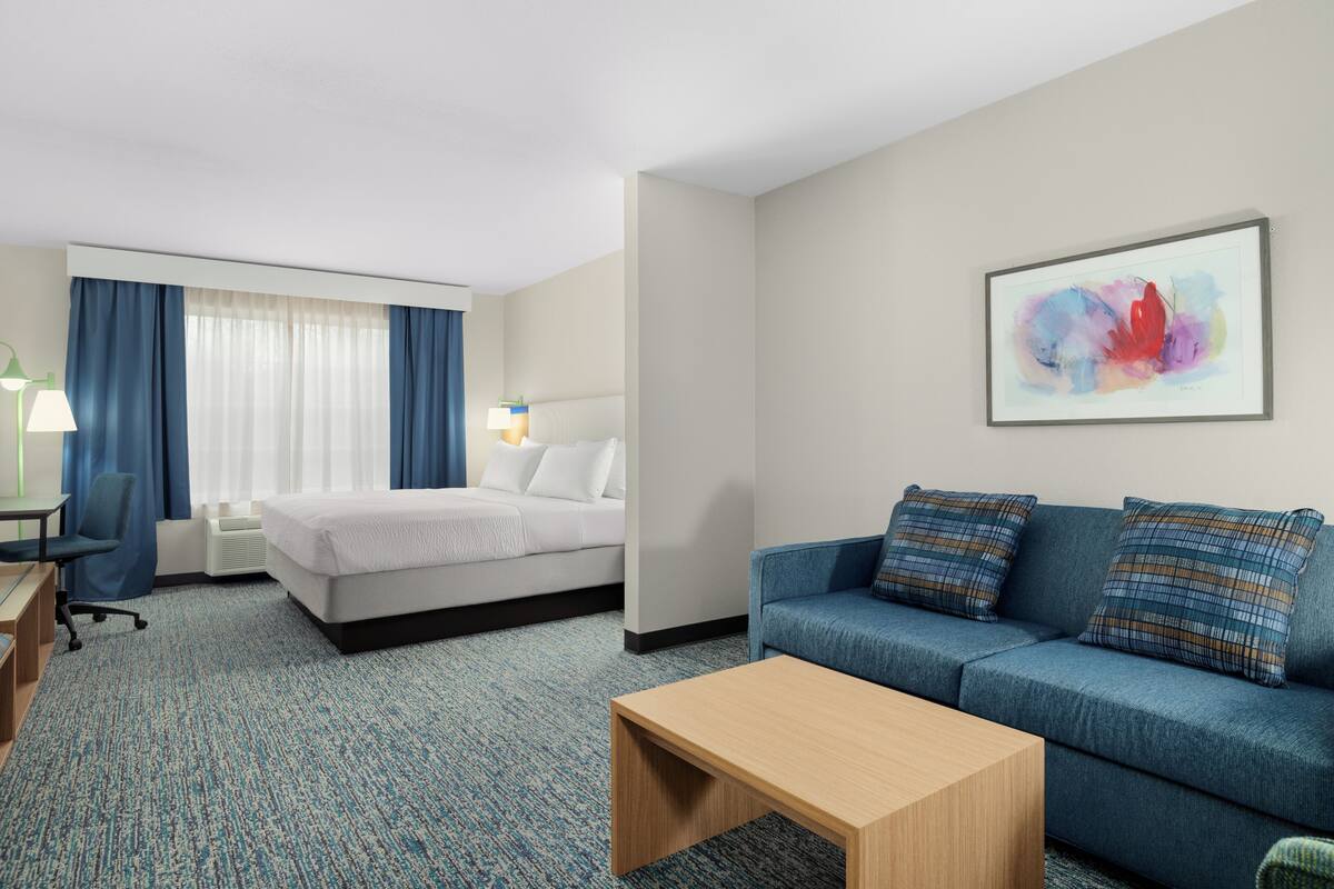 A hotel room with a bed, a blue sofa, a wooden coffee table, a desk with a chair, and a painting on the wall.