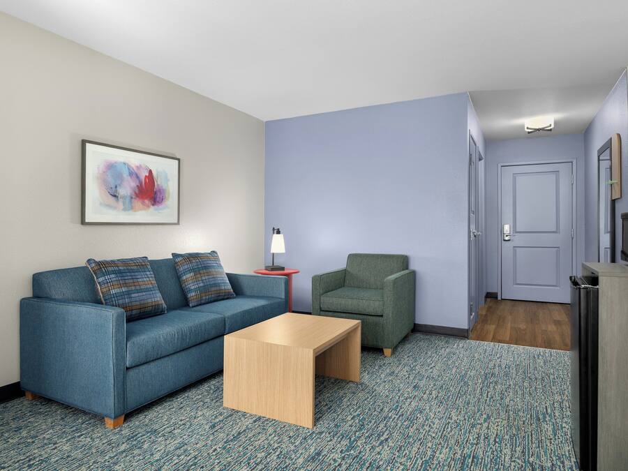 Una habitación con un sofá azul, un sillón verde, una mesa de centro de madera, un cuadro abstracto enmarcado, una mesita con una lámpara y una puerta que da a otra habitación.