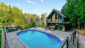 A modern house with a swimming pool, surrounded by a wooden deck and a forested area.