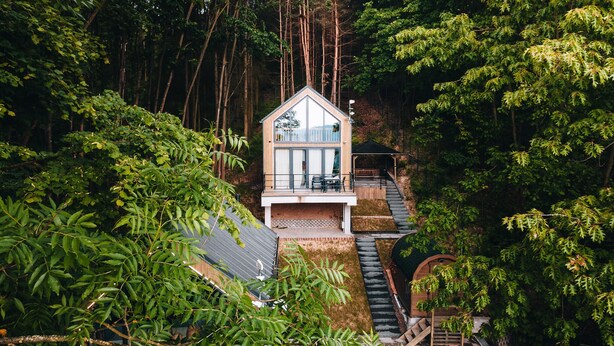 A modern house with a large glass window, surrounded by dense forest.