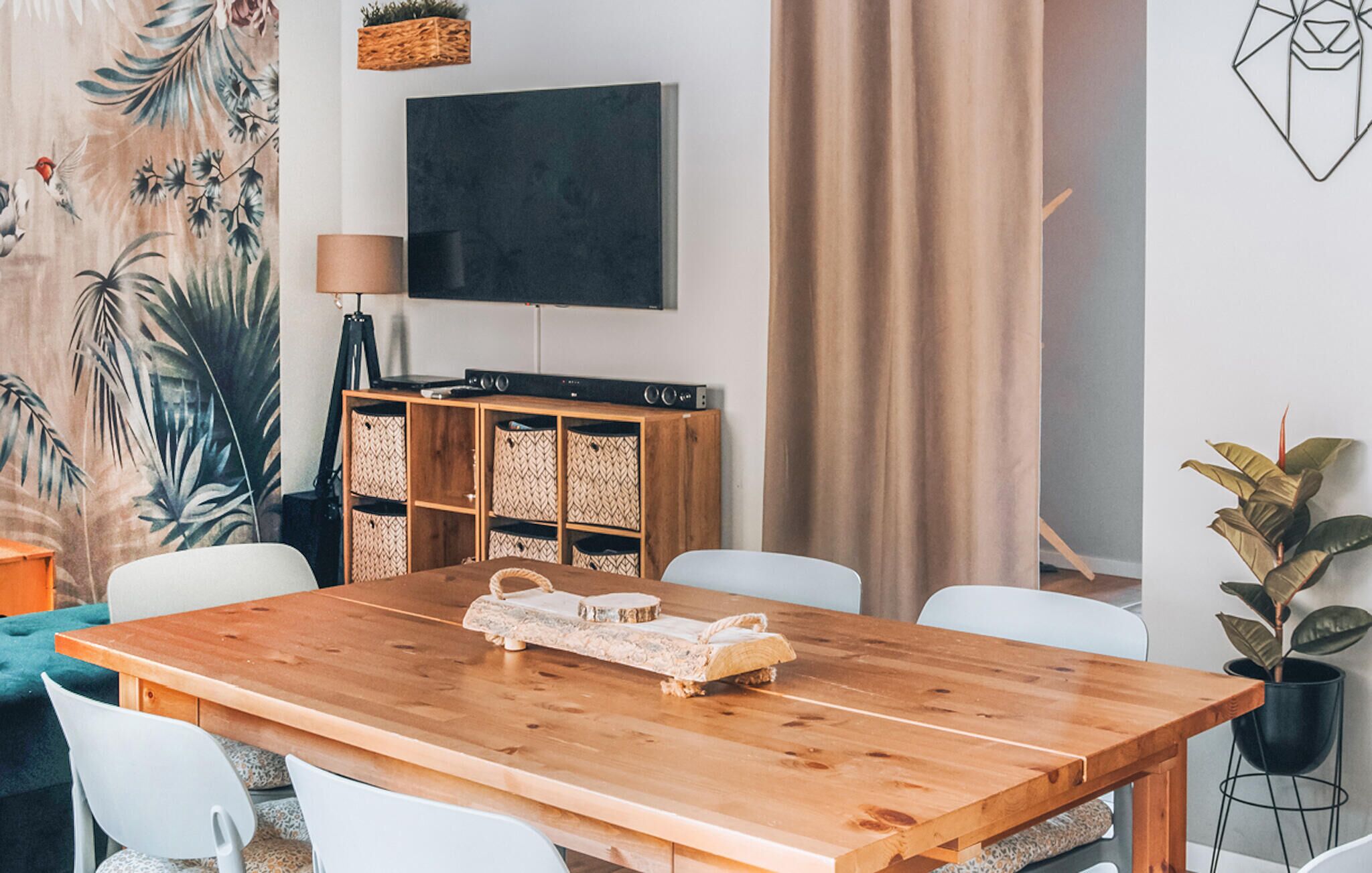 Una sala de estar moderna con una mesa de comedor de madera, sillas blancas, un televisor de pantalla plana, un sistema de sonido y una planta grande.