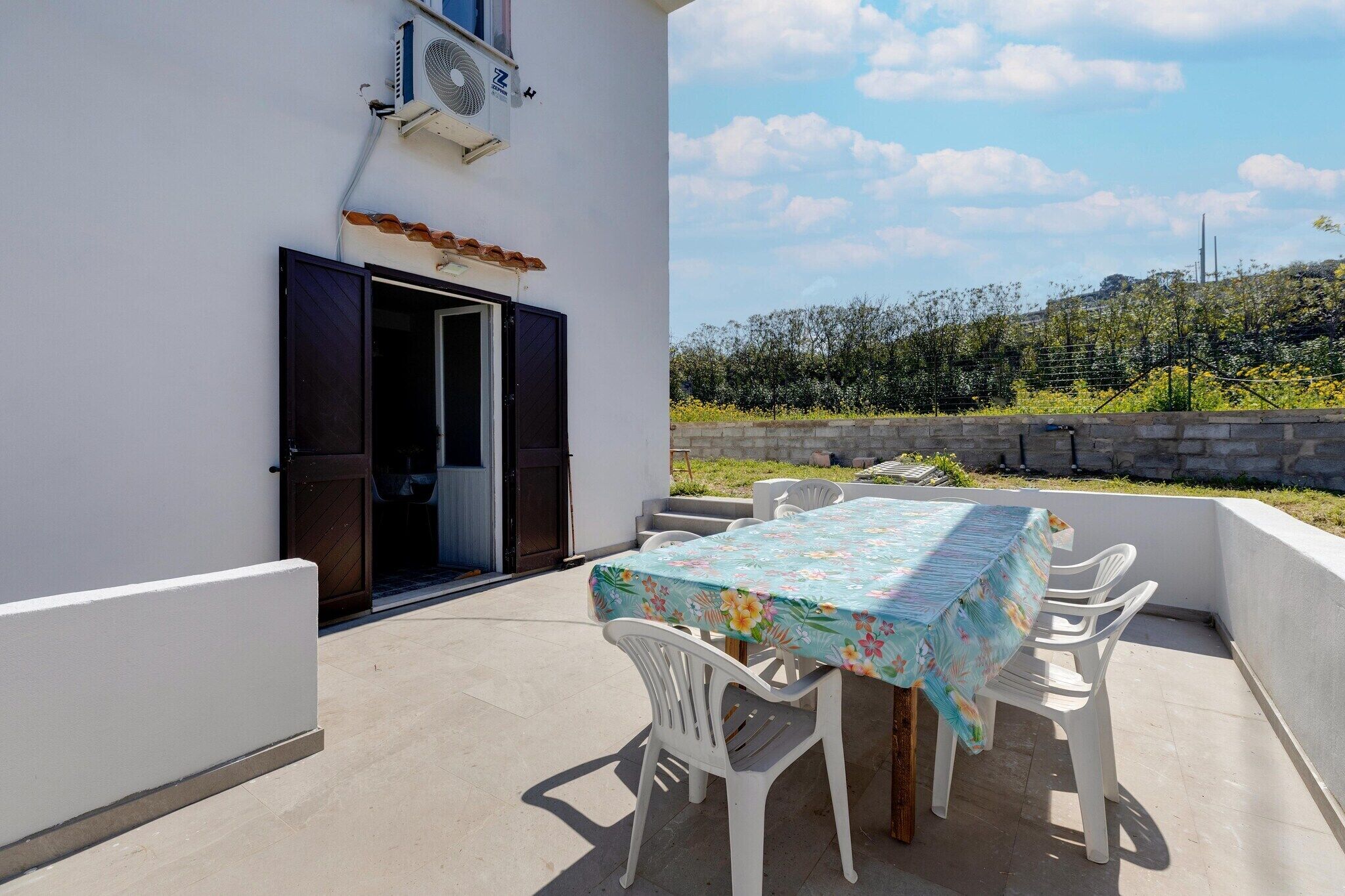 An outdoor patio with a table and chairs, a building entrance, and a view of a hill with vegetation.