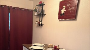 A dining area with a table set for four, including plates, cutlery, and glasses. There are two chairs with cushioned seats. A red curtain is partially visible on the left, and a wall-mounted shelf with decorative items is seen above the table.