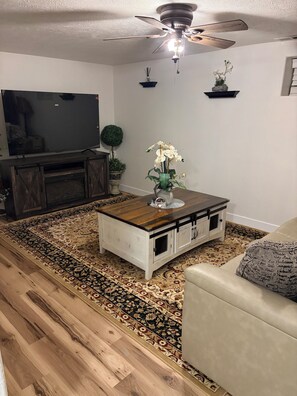 Una sala de estar con una mesa de centro de madera, un sofá color beige, un televisor y un ventilador de techo.