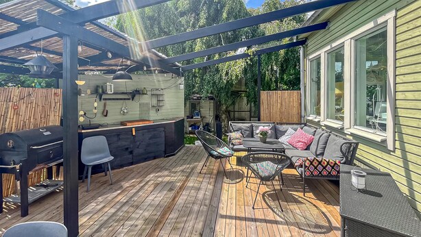 Eine überdachte Terrasse mit Grill, Sitzgelegenheiten und Blick auf das Haus.