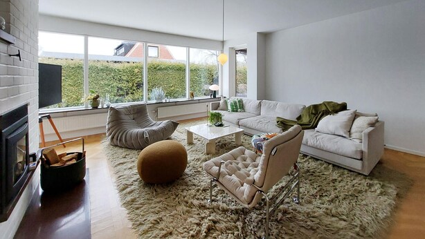 Un salon moderne comprenant un canapé, un fauteuil, un pouf et une table basse. On y trouve également une cheminée, une grande fenêtre donnant sur un paysage verdoyant et une lampe.