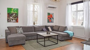 A modern living room with a grey sectional sofa, a small coffee table, and two windows with white curtains. There are two wall-mounted air conditioners and a chandelier. - Cozy home in Gärds Köpinge with WiFi (Gärds Köpinge)