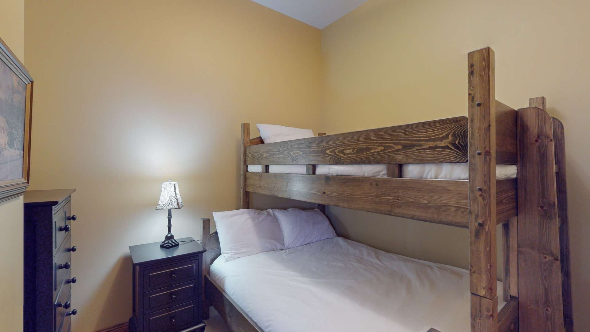A small, single-bed room with a wooden headboard, a bedside table with a lamp, and a dresser with multiple drawers.