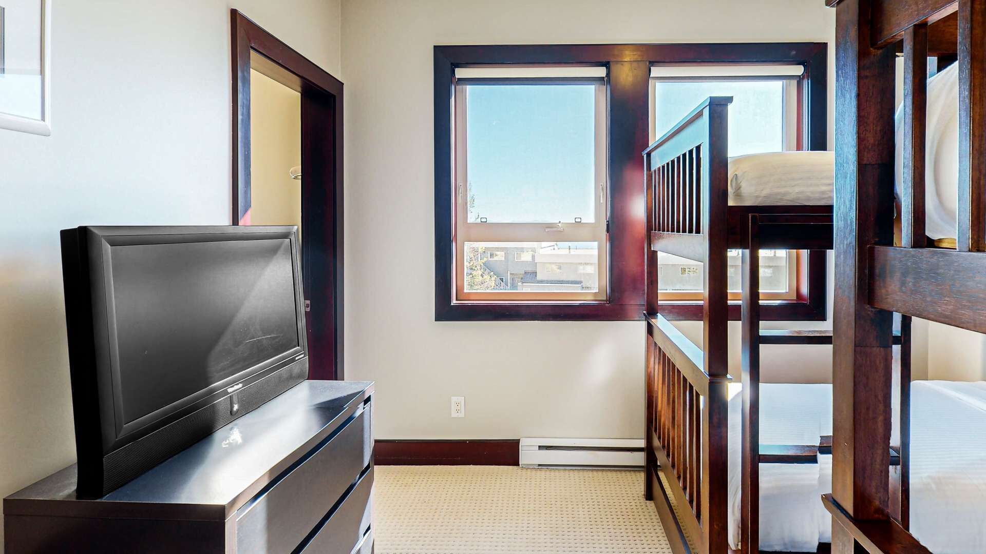 Une petite chambre comprenant un lit, une télévision et une fenêtre donnant sur des immeubles.