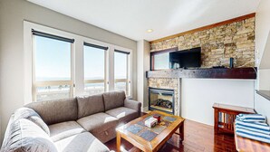 A living room with a sofa, a coffee table, a fireplace, a TV, and large windows. - Okanagan - Aspens Townhouse 1 (Big White Mountain)