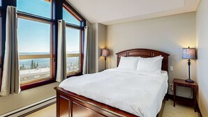 A hotel room with a large bed, two bedside lamps, and a view of a cityscape through large windows. - Okanagan - Aspens Townhouse 1 (Big White Mountain)