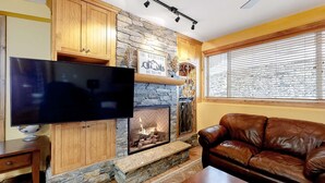 A living room with a stone fireplace, a flat-screen TV, wooden cabinets, and a leather couch. - Okanagan - Aspens 4307 (Big White Mountain)