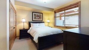 Een slaapkamer met een bed, twee nachtkastjes met lampen, een bijzettafeltje, een houten garderobe, een raam met jaloezieën en een lijst met een afbeelding aan de muur.