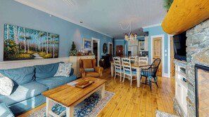 A living room with a sofa, coffee table, dining table with chairs, a fireplace, and a painting on the wall. - Okanagan - Aspens 4607 (Big White Mountain)